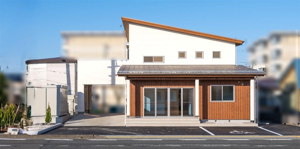 片流れ屋根の白い外壁に、木の縦張りの平屋部分が付いた福祉施設の外観。前面に駐車場
